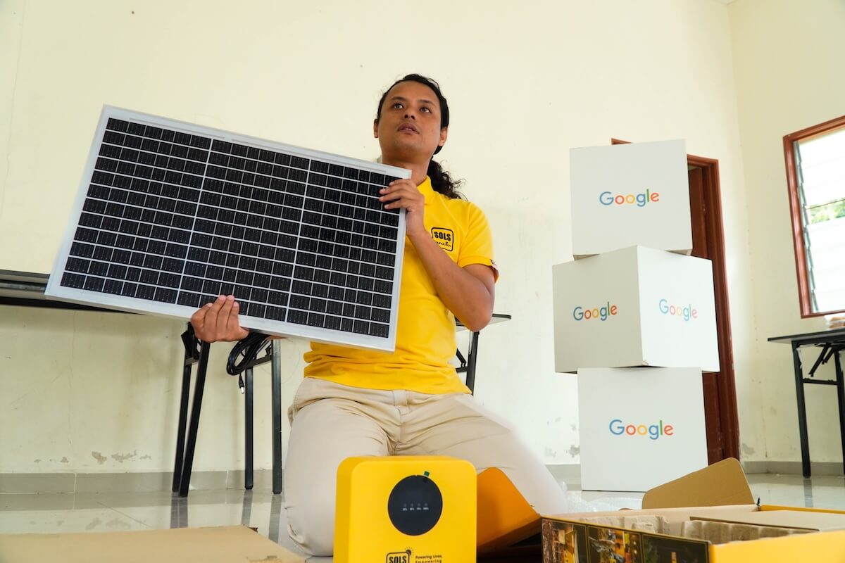 A SOLS Foundation team member holds a solar panel beside boxed home solar system components before installation.
