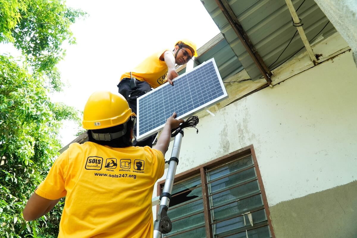 Two SOLS Foundation team members in hard hats mount a solar panel on a house roof.